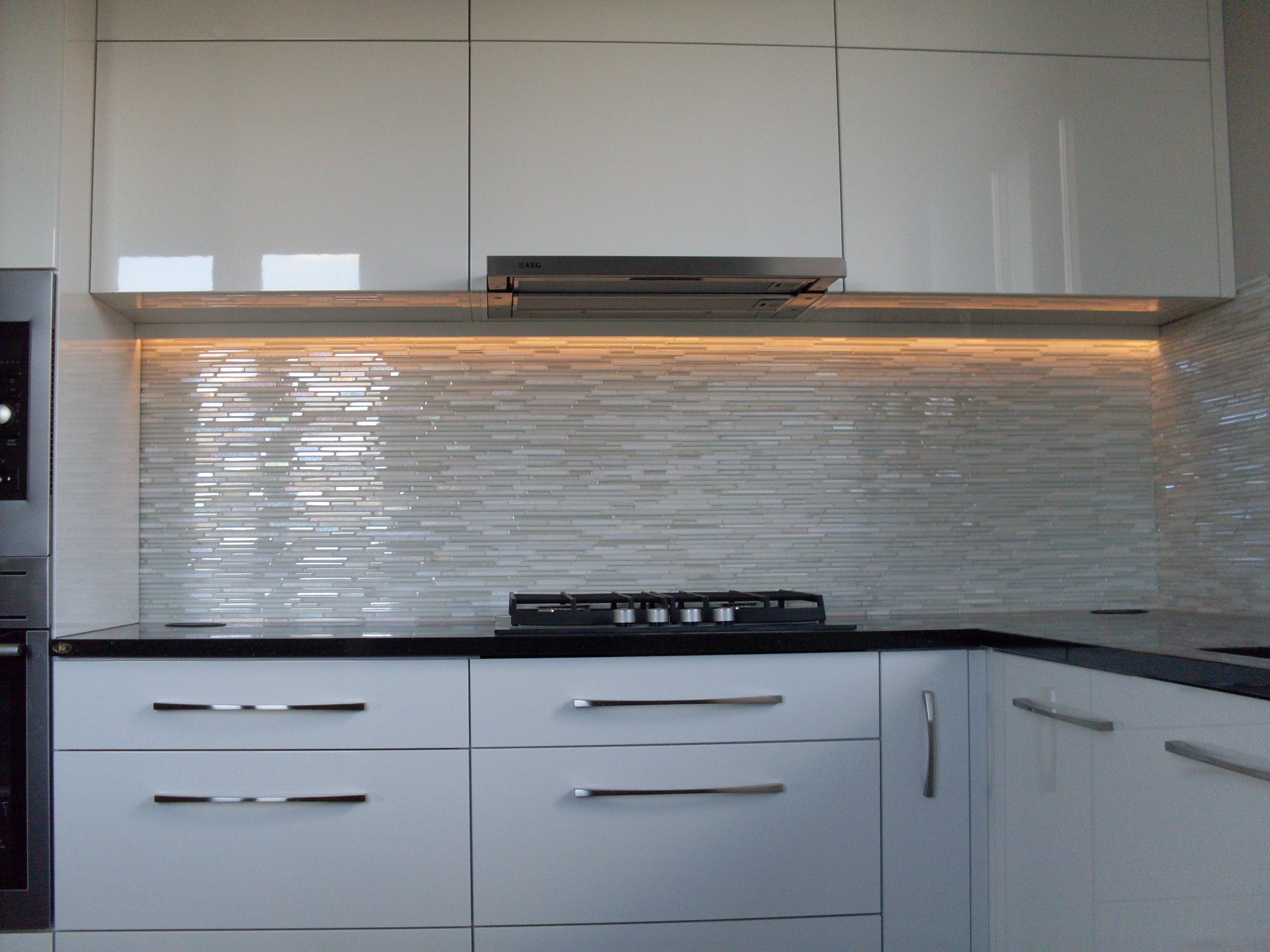 Biała, nowoczesna kuchnia z czarnym blatem, płytą gazową i mozaiką szklaną podświetloną ledami, widoczny okap marki AEG.