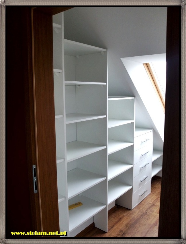 Biała zabudowa meblowa na wymiar w skosach poddasza, z półkami i szufladami, widoczna poziomica na jednej z półek, drewniana podłoga.