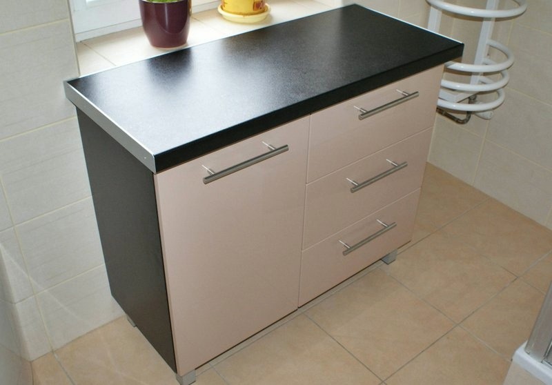 Szafka łazienkowa z trzema szufladami i dwojgiem drzwiczek, z czarnym blatem i srebrnymi uchwytami, na tle kremowych płytek i grzejnika drabinkowego.