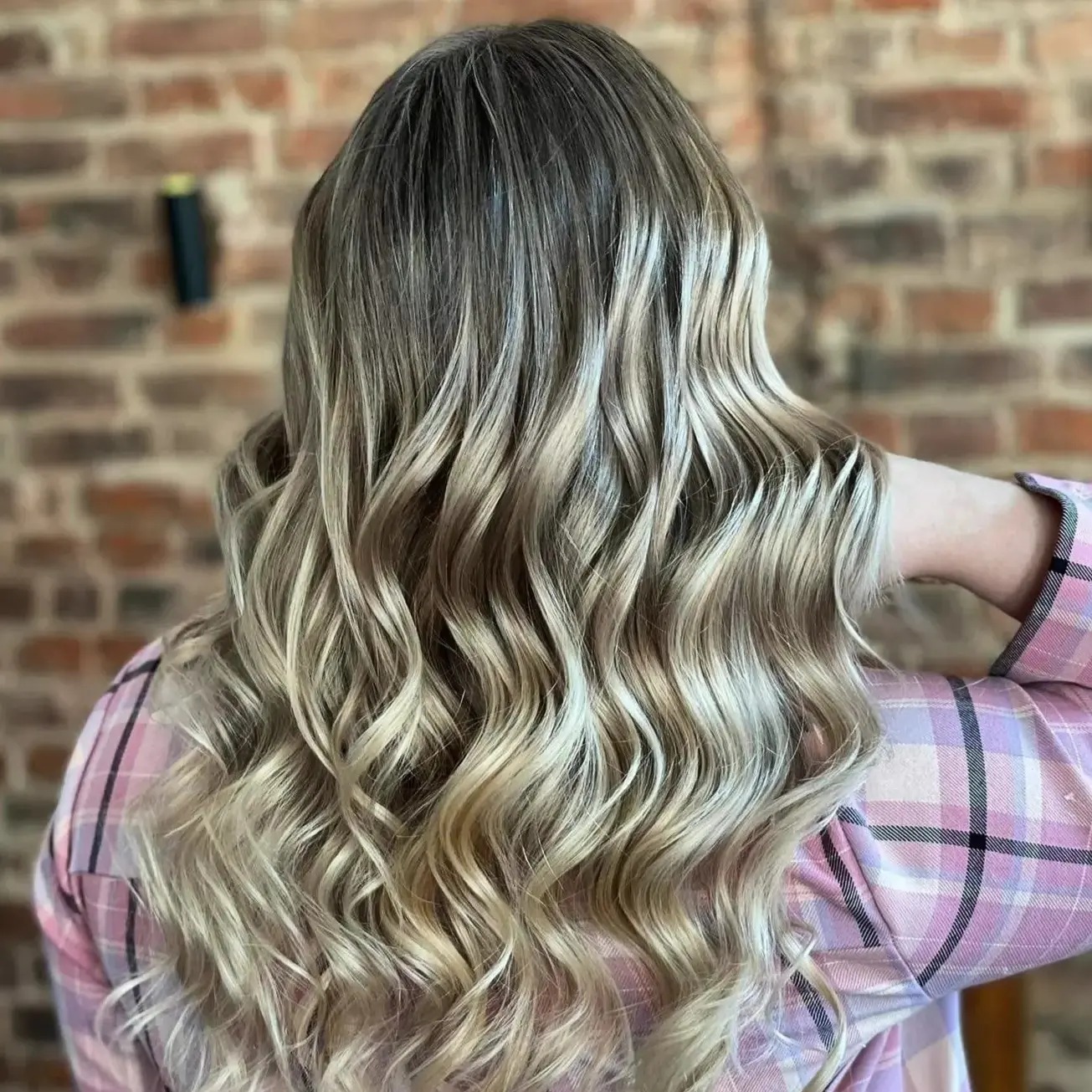Długie, falowane włosy z balejażem w odcieniach blondu i brązu, na tle ceglanej ściany, widoczne ramię w różowej koszuli w kratę.