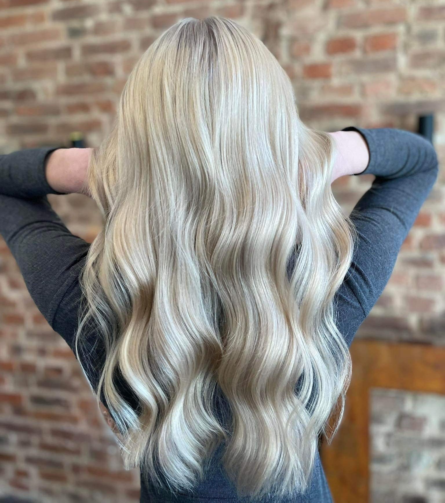 Długie, platynowe blond włosy z delikatnymi falami, widoczne od tyłu, z ciemniejszymi odrostami, na tle ceglanej ściany.