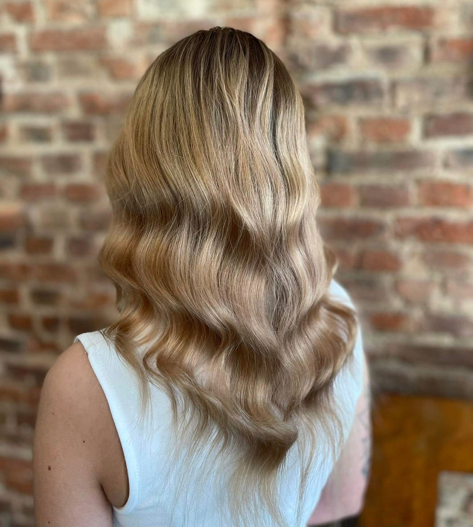 Tył głowy kobiety z długimi, falowanymi włosami w odcieniach blondu i brązu. Fryzura wykonana w Mierzynie na tle ściany z czerwonej cegły.