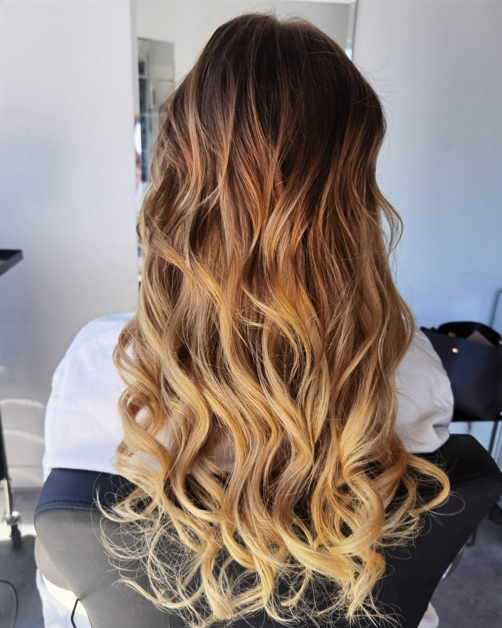 Długie, falowane włosy z ombre, od ciemnego brązu u nasady do jasnego blondu na końcach, widok z tyłu w salonie.