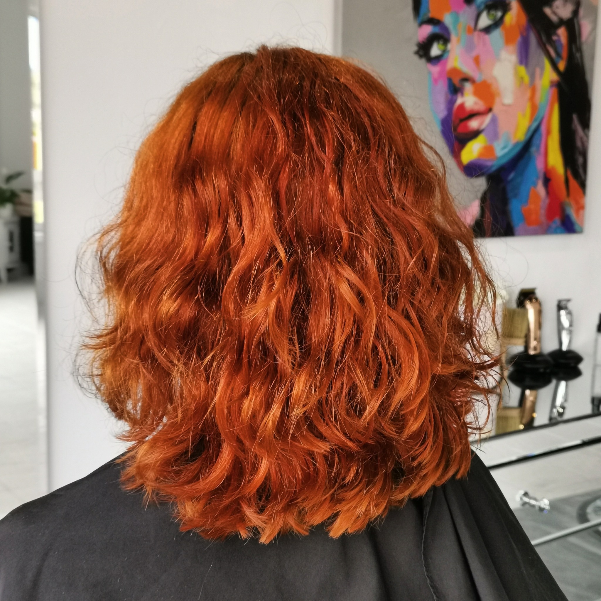 Widok z tyłu na krótkie, falowane, intensywnie rude włosy klientki w salonie, z widocznym czarnym okryciem ochronnym i fragmentem kolorowego obrazu na ścianie.