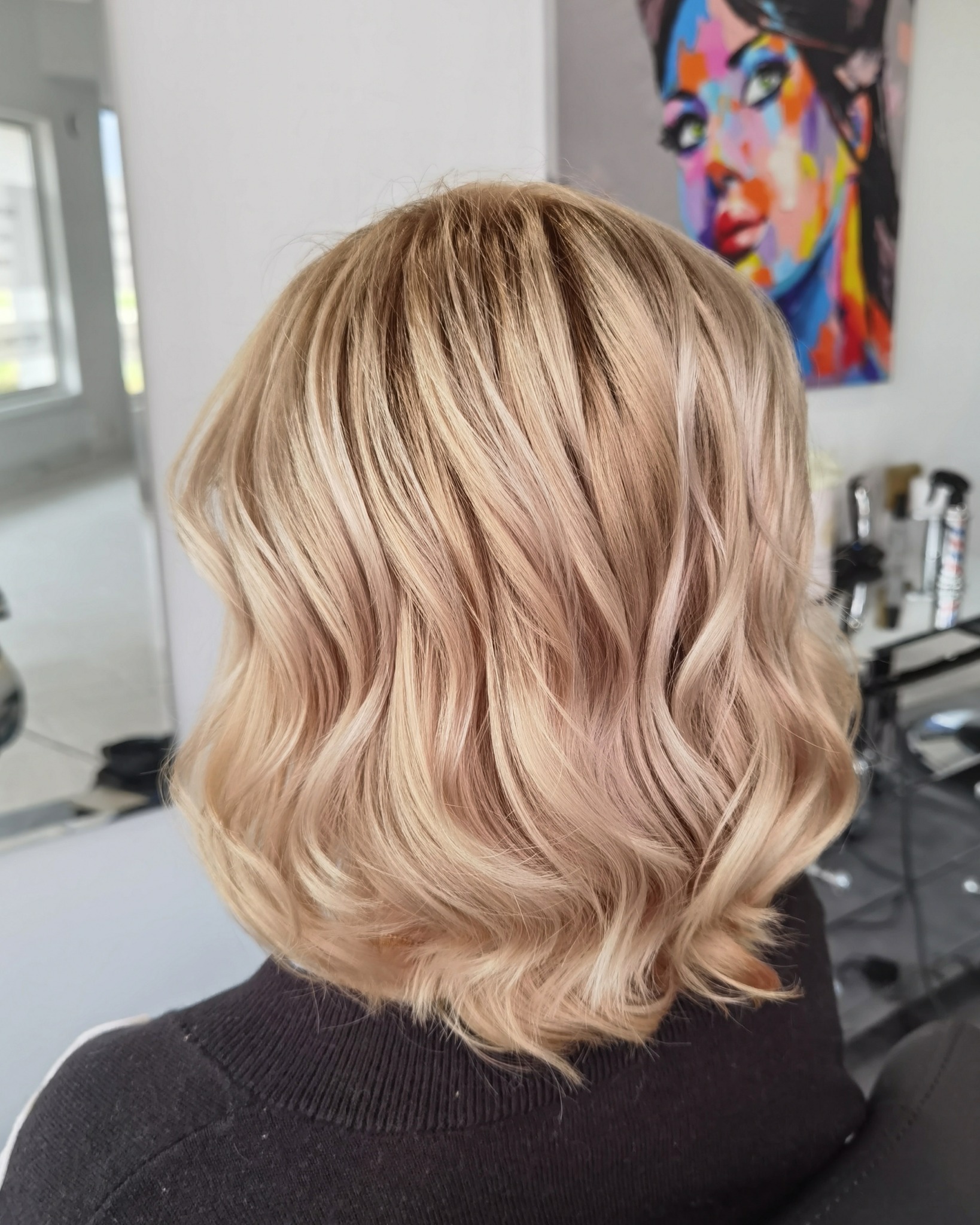 Krótkie, falowane włosy w odcieniach blondu, widok z tyłu, z subtelnymi refleksami i kolorowym portretem w tle.