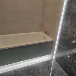 Niedokończona obudowa wanny z płyt gipsowo-kartonowych, widoczne przyłącza wodne i grzejnik drabinkowy na tle szarych płytek imitujących kamień.