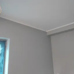 Szara ściana po malowaniu z widoczną białą listwą przypodłogową i ramą okienną z folią ochronną.