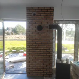 Ściana wyłożona cegłą klinkierową z dwoma otworami na rury, widoczny kominek i rura odprowadzająca dym, przez okno widok na ogród w słoneczny dzień.