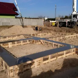 Wylane fundamenty domu jednorodzinnego z drewnianym szalunkiem, widoczna pompa do betonu i dwóch pracowników w tle.