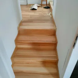 Jasne, drewniane schody z widocznymi śladami taśmy malarskiej na krawędziach, prowadzące do pomieszczenia z drewnianą podłogą, na kt&oacute;rej stoi krzesło z rośliną doniczkową i leżą klapki.