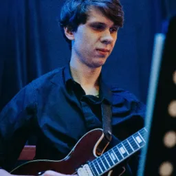 Młody mężczyzna w czarnej koszuli gra na gitarze elektrycznej typu hollow body na tle ciemnoniebieskiej zasłony. Widoczny fragment pulpitu do nut po prawej stronie.