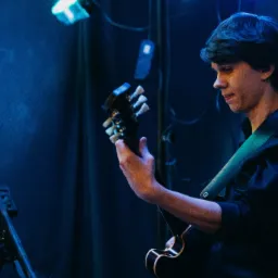 Młody muzyk w ciemnej koszuli, zamyślony, gra na gitarze elektrycznej na scenie oświetlonej niebieskim światłem, widoczny statyw na nuty.