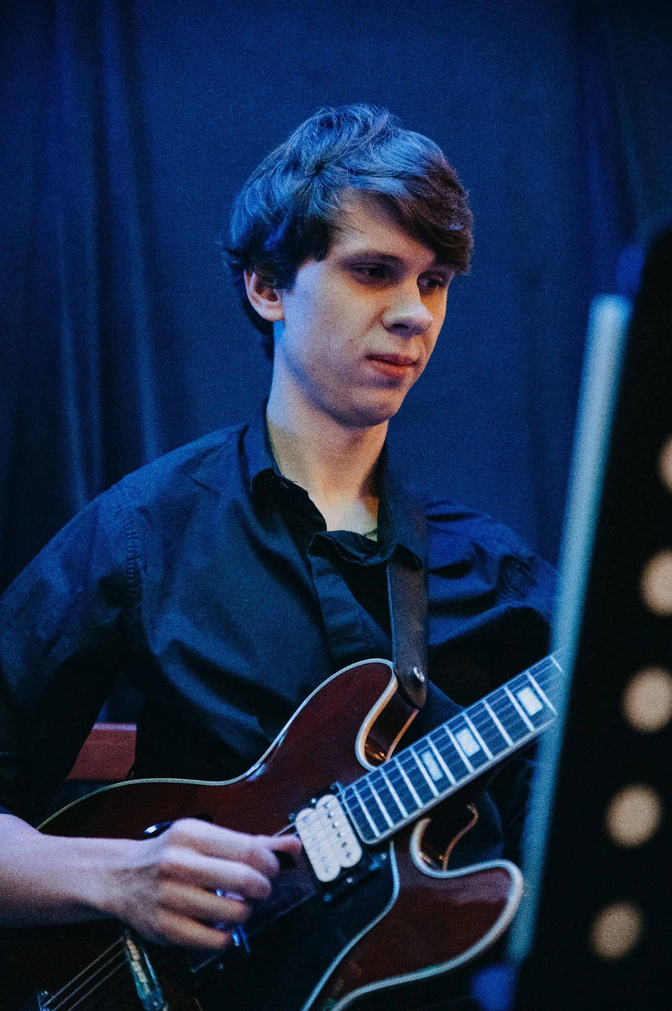 Młody mężczyzna w czarnej koszuli gra na gitarze elektrycznej typu hollow body na tle ciemnoniebieskiej zasłony. Widoczny fragment pulpitu do nut po prawej stronie.