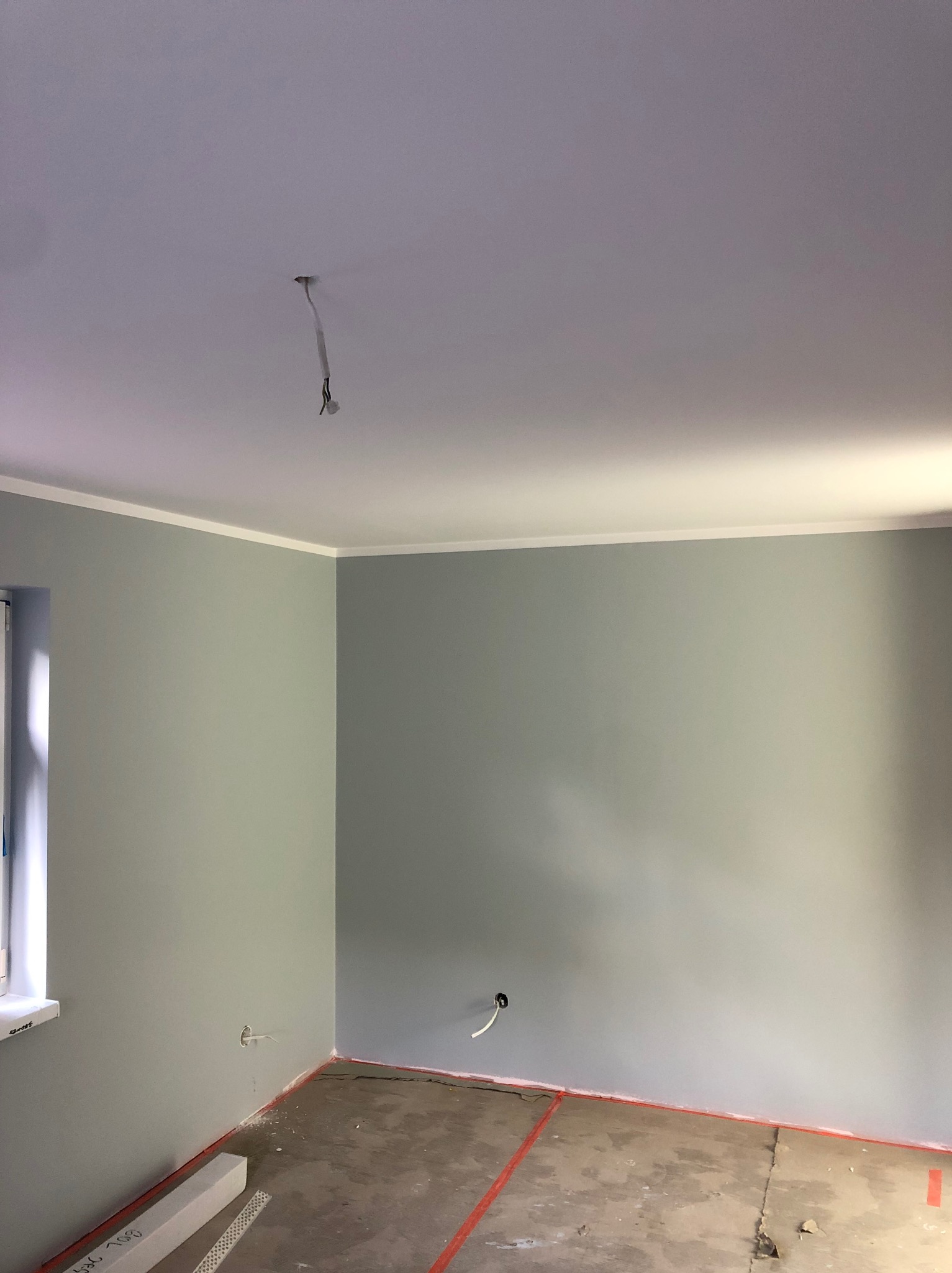 Pomieszczenie po malowaniu ścian na szaro, widoczne listwy przypodłogowe, sufit z listwą ozdobną i wystające kable. Podłoga przygotowana do dalszych prac.