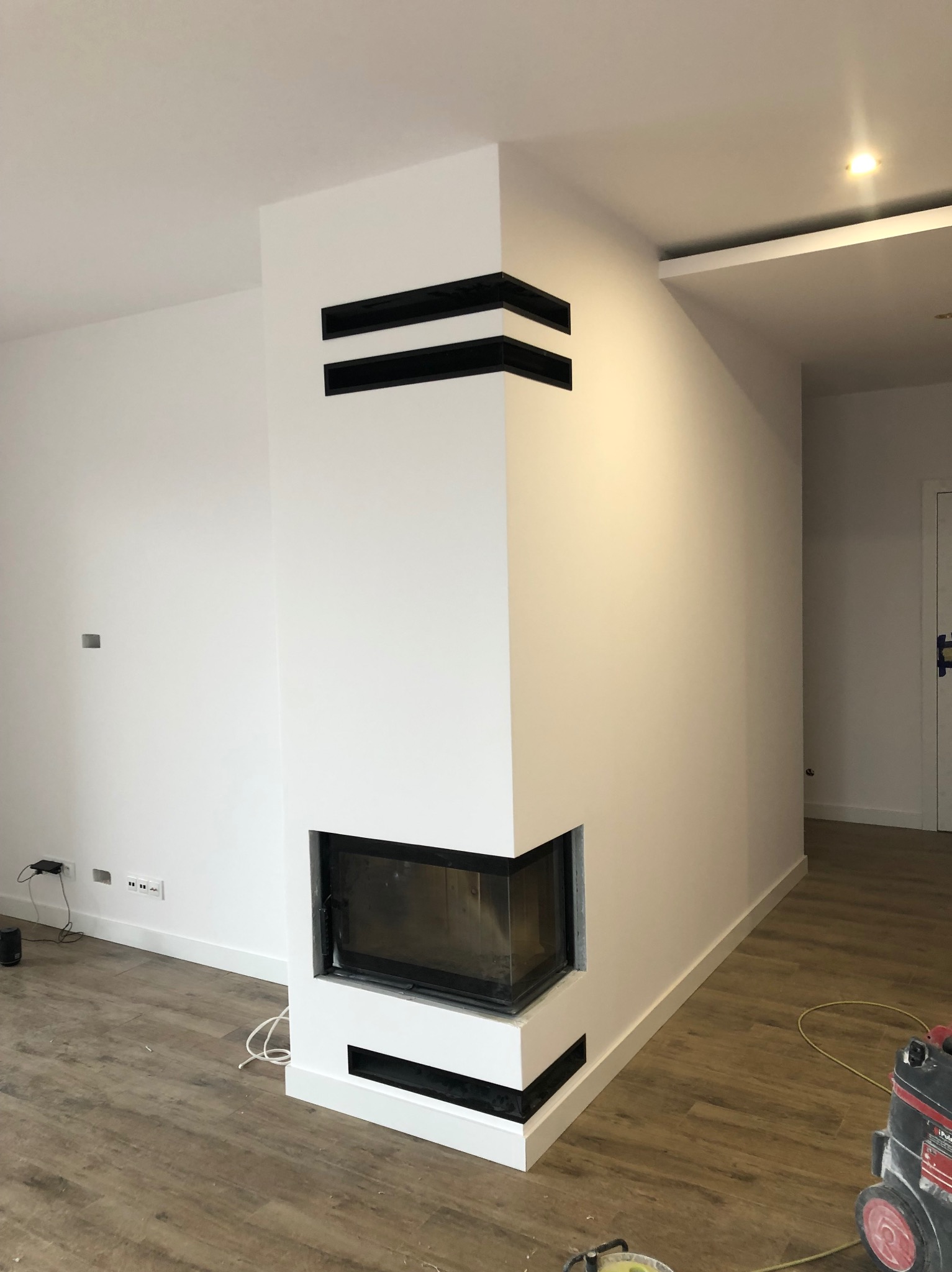 Biały kominek narożny z czarnymi akcentami w nowoczesnym salonie. Widoczne listwy przypodłogowe i drewnopodobna podłoga. Częściowo widoczny odkurzacz.