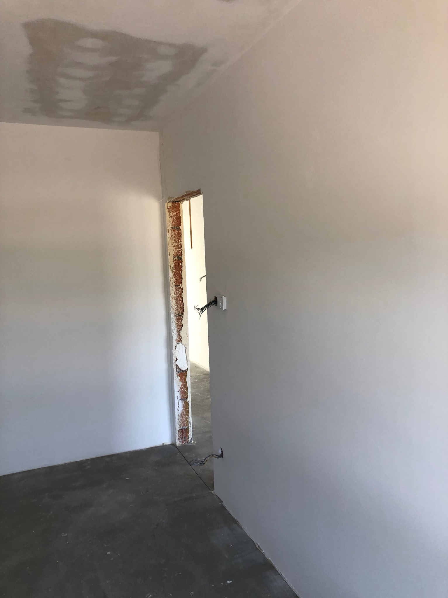 Wnętrze w trakcie remontu: świeżo wyszpachlowane ściany w szarym odcieniu, surowy beton na podłodze, widoczne przewody elektryczne i ościeżnica z fragmentami starej cegły.