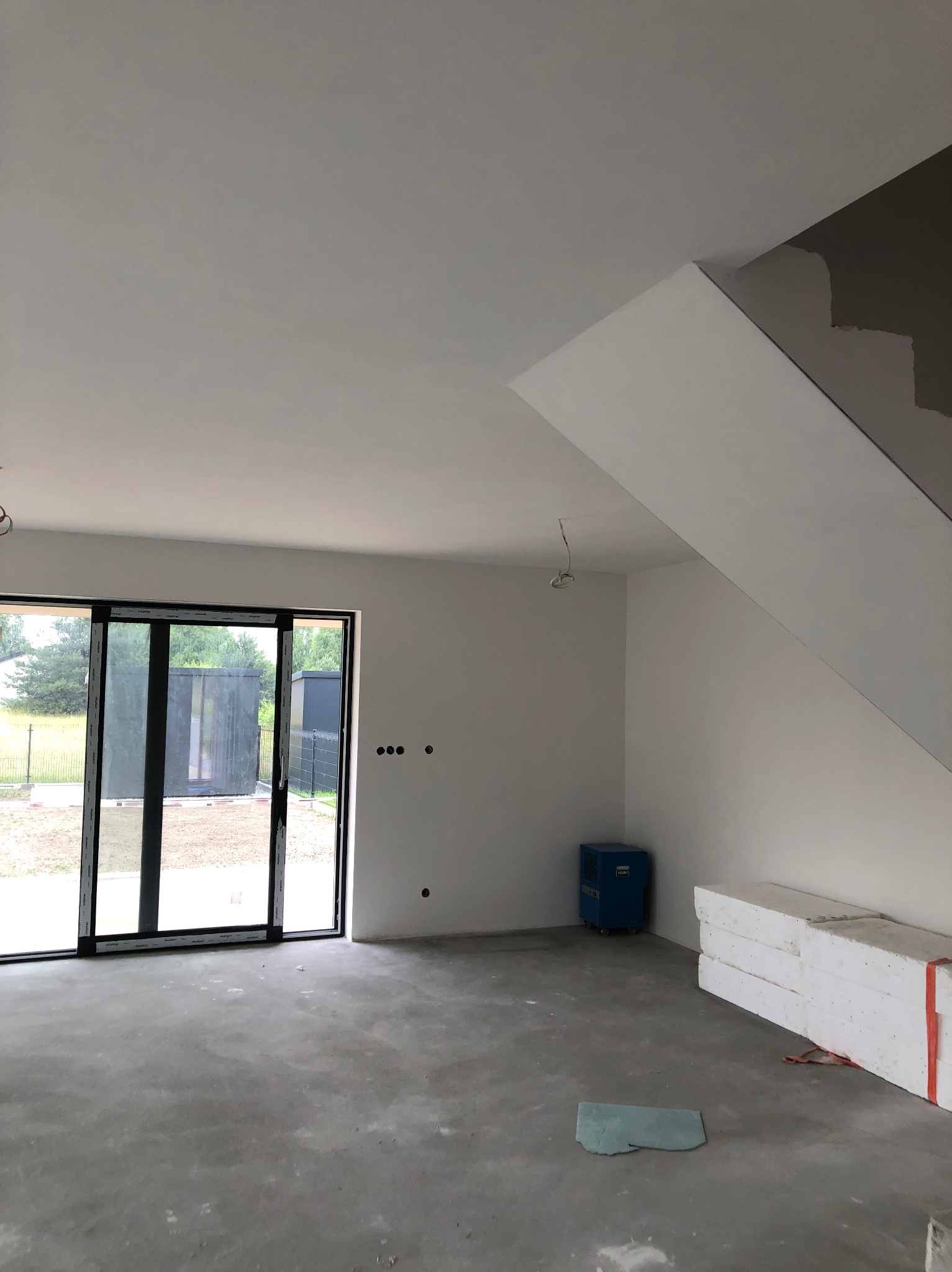 Surowe wnętrze domu w trakcie wykańczania: gładkie, białe ściany, betonowa podłoga, duże okno przesuwne, widoczne materiały budowlane i osuszacz powietrza.