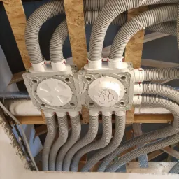 Widok z dołu na rozdzielacz rekuperacji z podłączonymi elastycznymi przewodami wentylacyjnymi, zamontowany na drewnianej konstrukcji stropu, jeden z rozdzielaczy oznaczony napisem na białej naklejce.