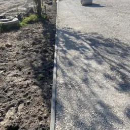 Walec drogowy ubija świeżo wysypaną warstwę kruszywa na przygotowywanej drodze, widoczny obok pas ziemi z obrzeżem i drzewem, w tle krajobraz polny.