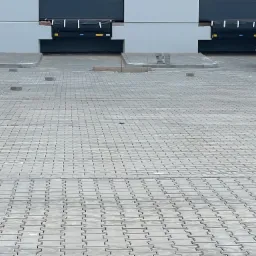 Układanie szarej kostki brukowej typu 'T' przed budynkiem magazynowym z rampami, widoczne palety z kostką i pojedyncze kostki przygotowane do ułożenia.