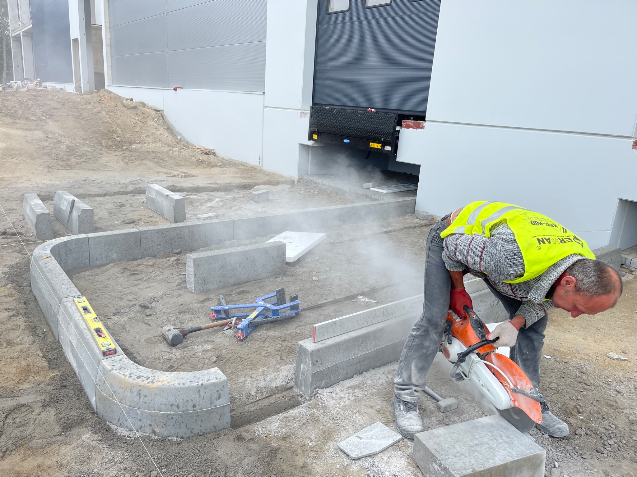 Pracownik w kamizelce odblaskowej przecina betonową obrzeże kostki brukowej za pomocą przecinarki Stihl, przygotowując ją do ułożenia przy rampie magazynu PANATONI.