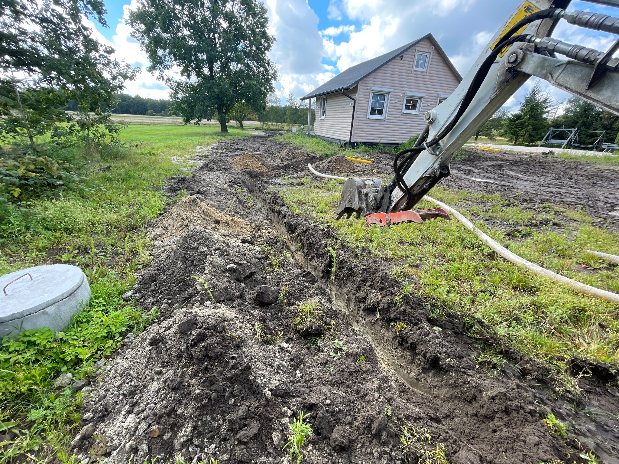 Wykonany wykop wąskoprzestrzenny pod rury na terenie posesji z domem w tle, widoczny fragment ramienia koparki i betonowy element studzienki.