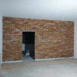 Ściana wykończona cegłą klinkierową w odcieniach czerwieni i brązu, widoczne przewody elektryczne wystające ze ściany, przygotowane do podłączenia oświetlenia, odkurzacz przemysłowy w przejściu...