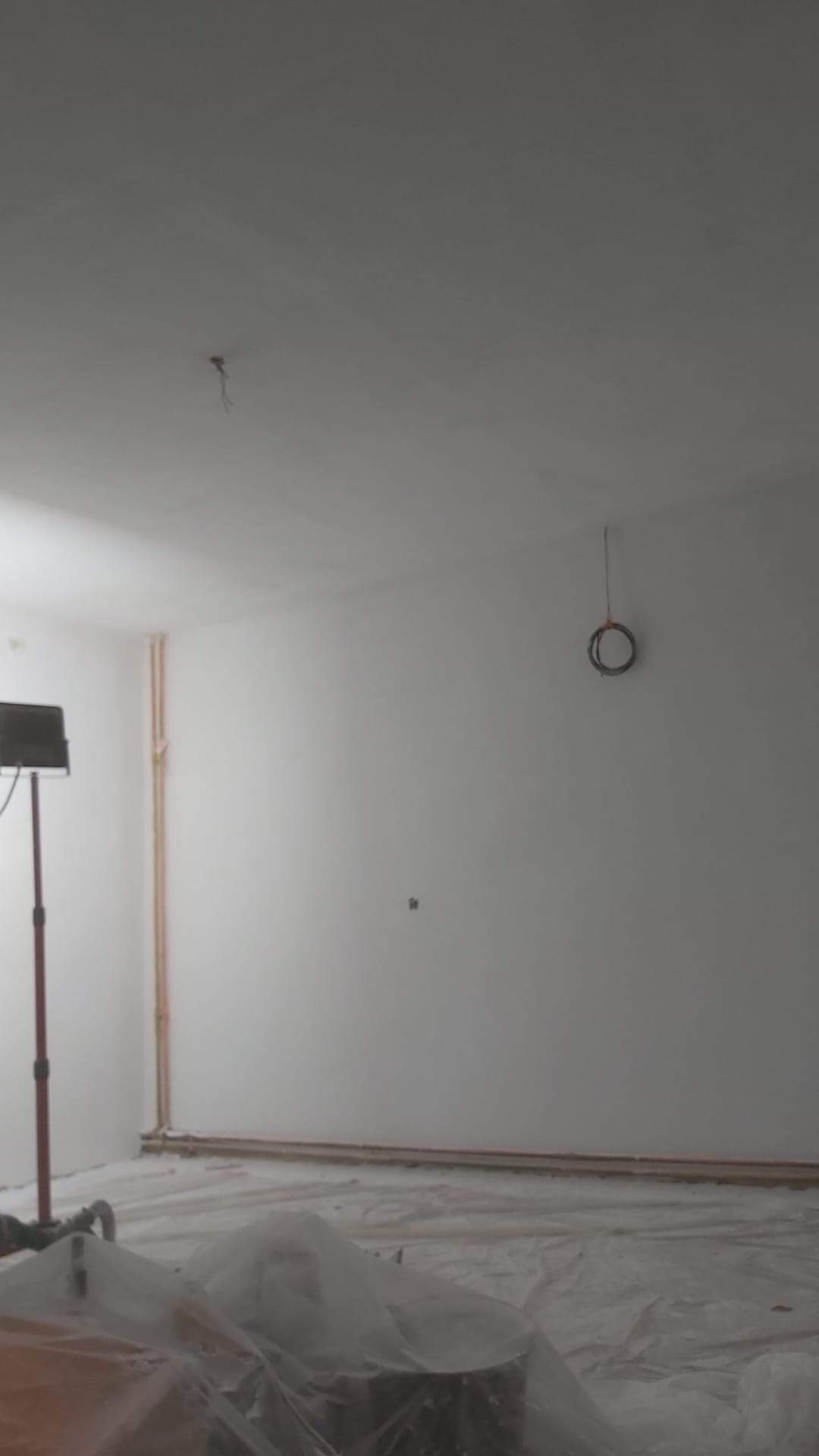 Przygotowane pomieszczenie do prac wykończeniowych: zakryta folią podłoga, lampa budowlana i świeżo pomalowane ściany z widocznymi rurami.
