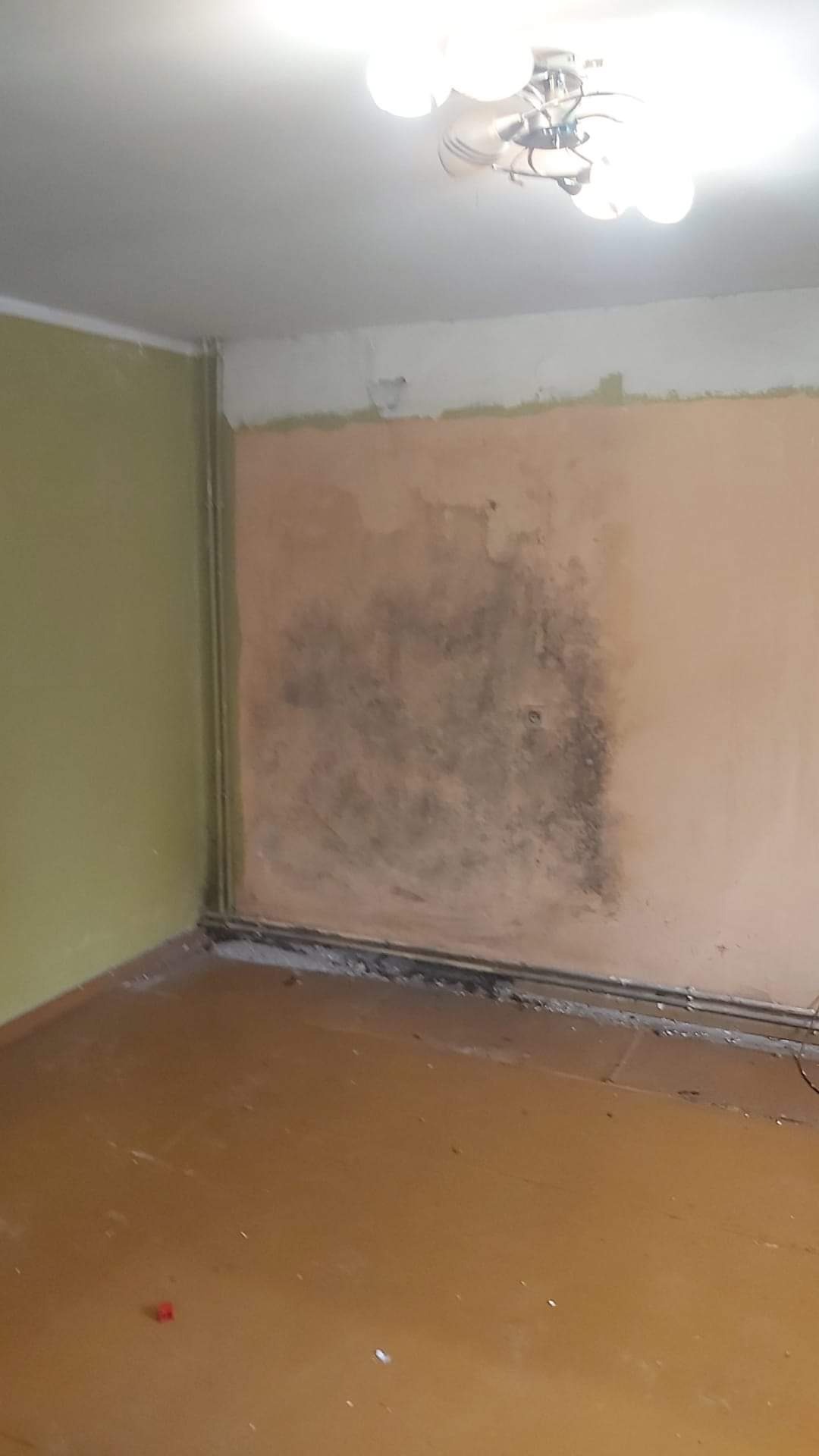 Pokój przed remontem: ściana z widocznym zawilgoceniem i pleśnią, odsłonięty fragment tynku, stara podłoga, widoczna instalacja grzewcza przy podłodze.