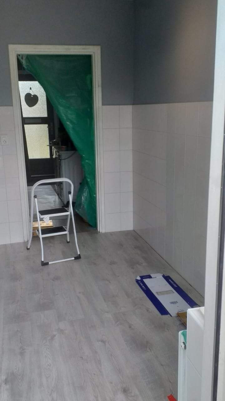 Wnętrze w trakcie prac wykończeniowych: zabezpieczone folią drzwi, drabinka malarska i częściowo położona podłoga z paneli w odcieniach szarości.