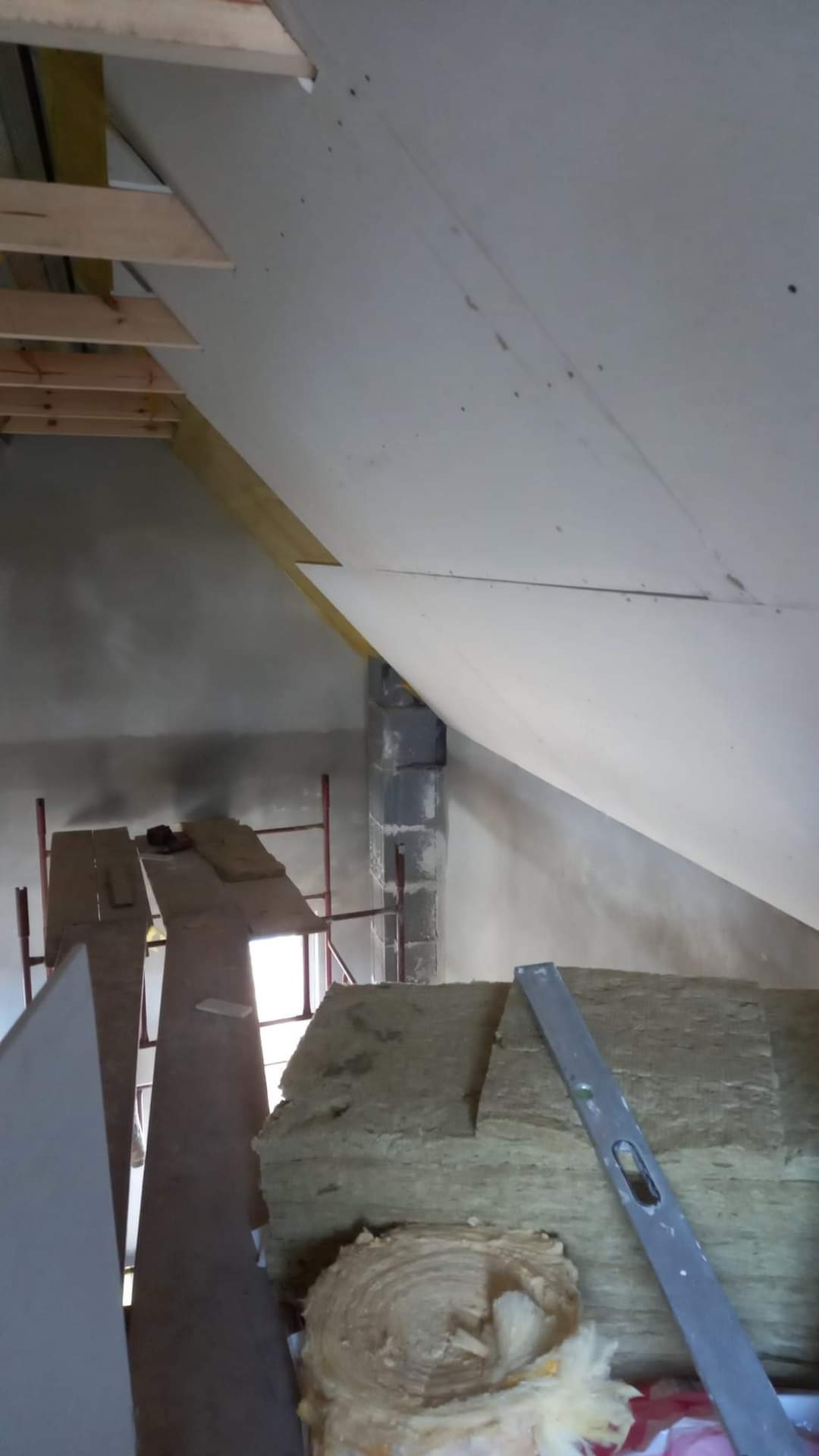 Poddasze w trakcie prac wykończeniowych: widoczna izolacja z wełny mineralnej, płyty gipsowo-kartonowe, rusztowanie i poziomica.