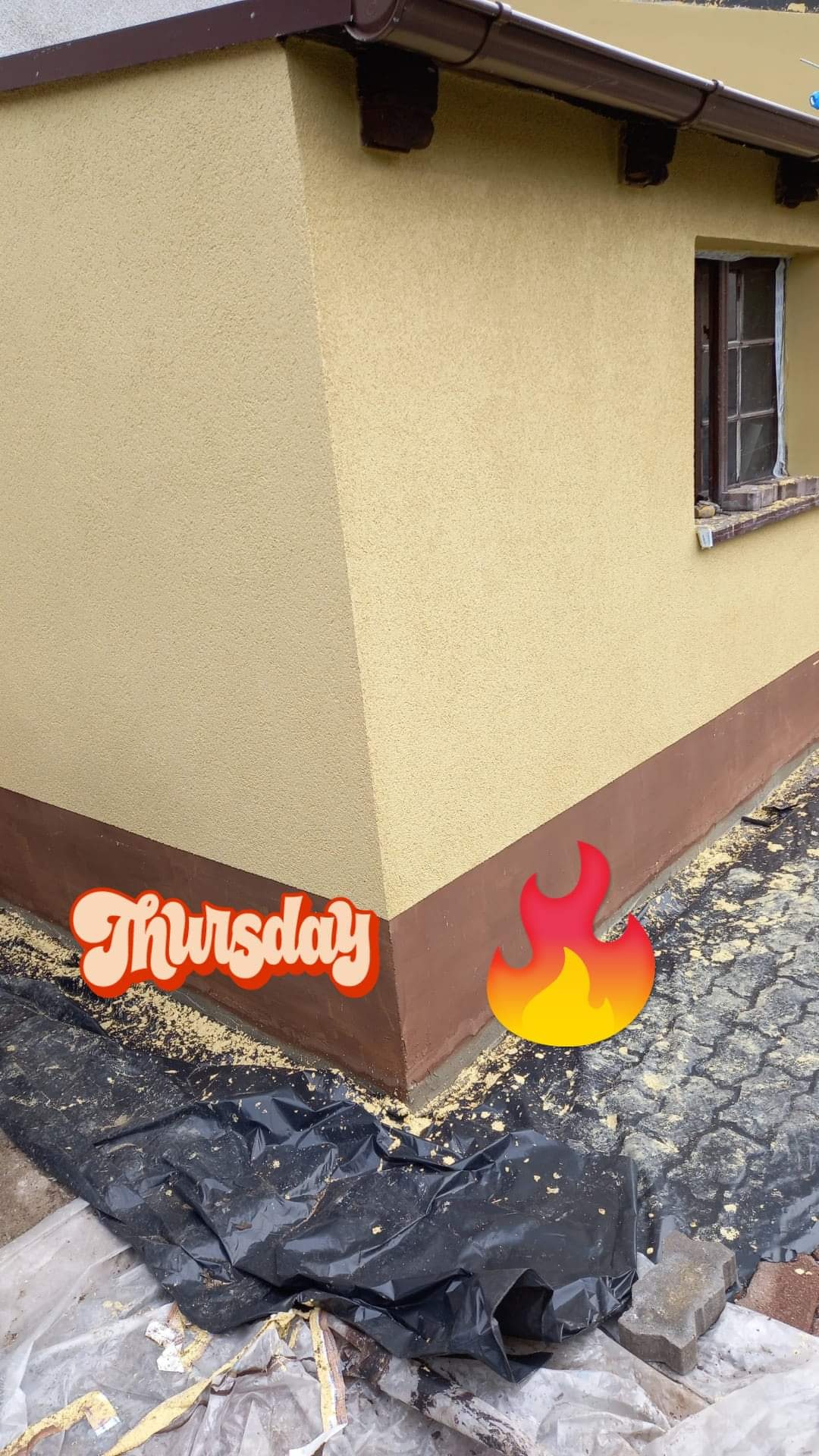 Fragment elewacji budynku pokrytej świeżym, beżowym tynkiem strukturalnym, z brązowym cokołem i rynną. Widoczne zabezpieczenia folią i resztki materiałów budowlanych na kostce brukowej.