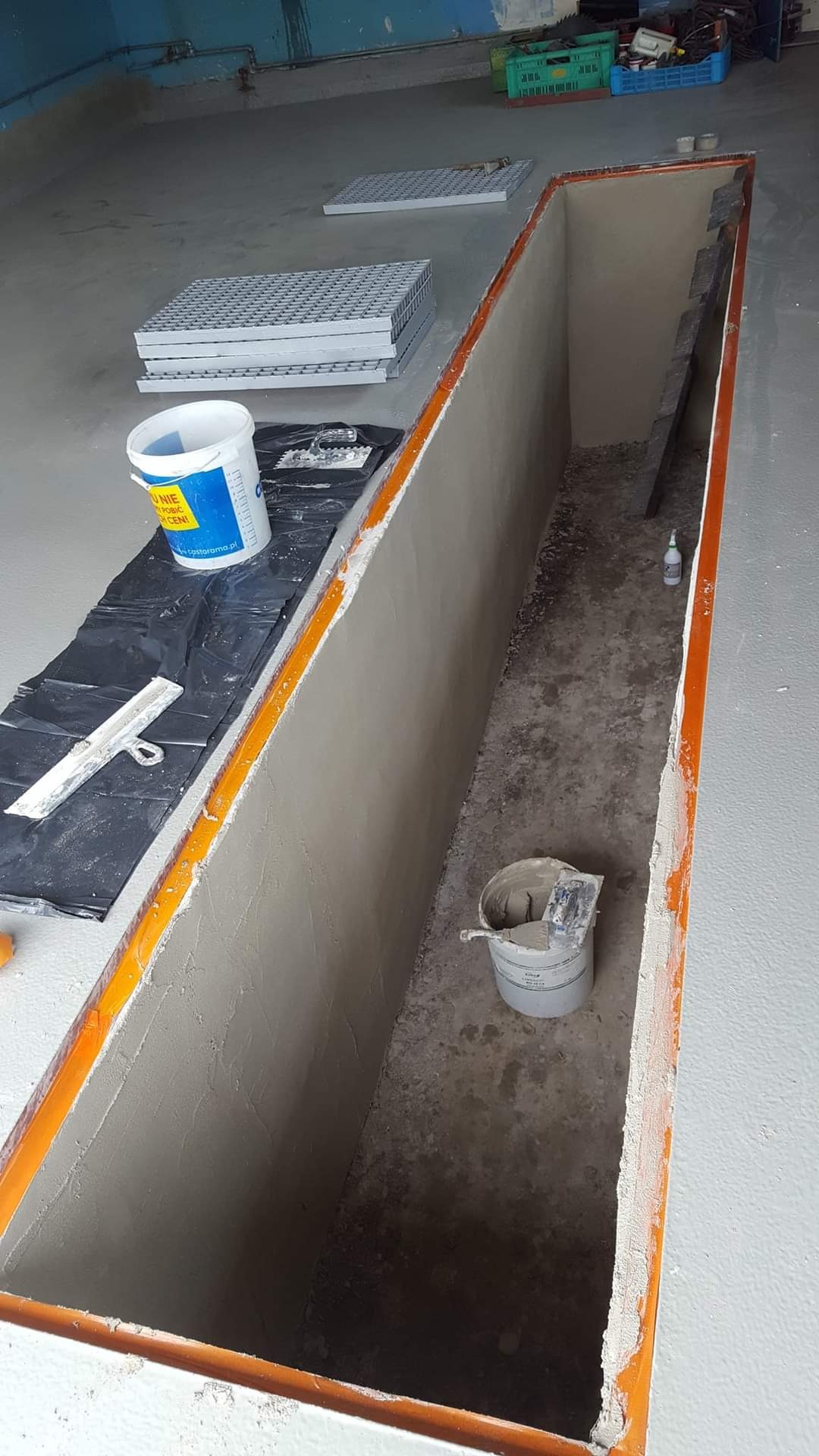 Wnętrze kanału warsztatowego w trakcie prac, ściany pokryte świeżym tynkiem, widoczne narzędzia i wiadro z zaprawą, kraty wentylacyjne obok.