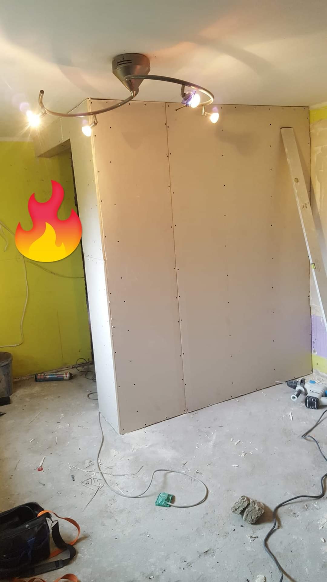 Wnętrze w trakcie remontu: ściana działowa z płyt gipsowo-kartonowych, widoczne narzędzia, surowy beton na podłodze, lampa sufitowa z kilkoma reflektorami, fragment pomalowanej na zielono ściany...