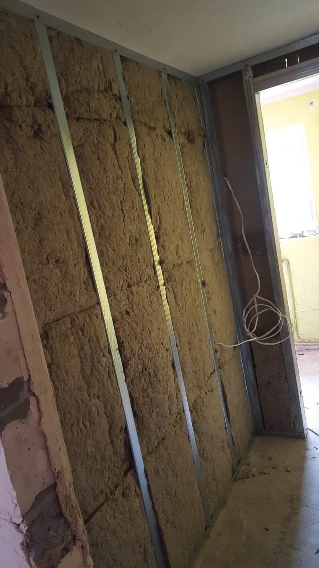 Ściana wewnętrzna w trakcie ocieplania wełną mineralną, z widocznymi metalowymi profilami konstrukcyjnymi i wystającymi przewodami elektrycznymi.