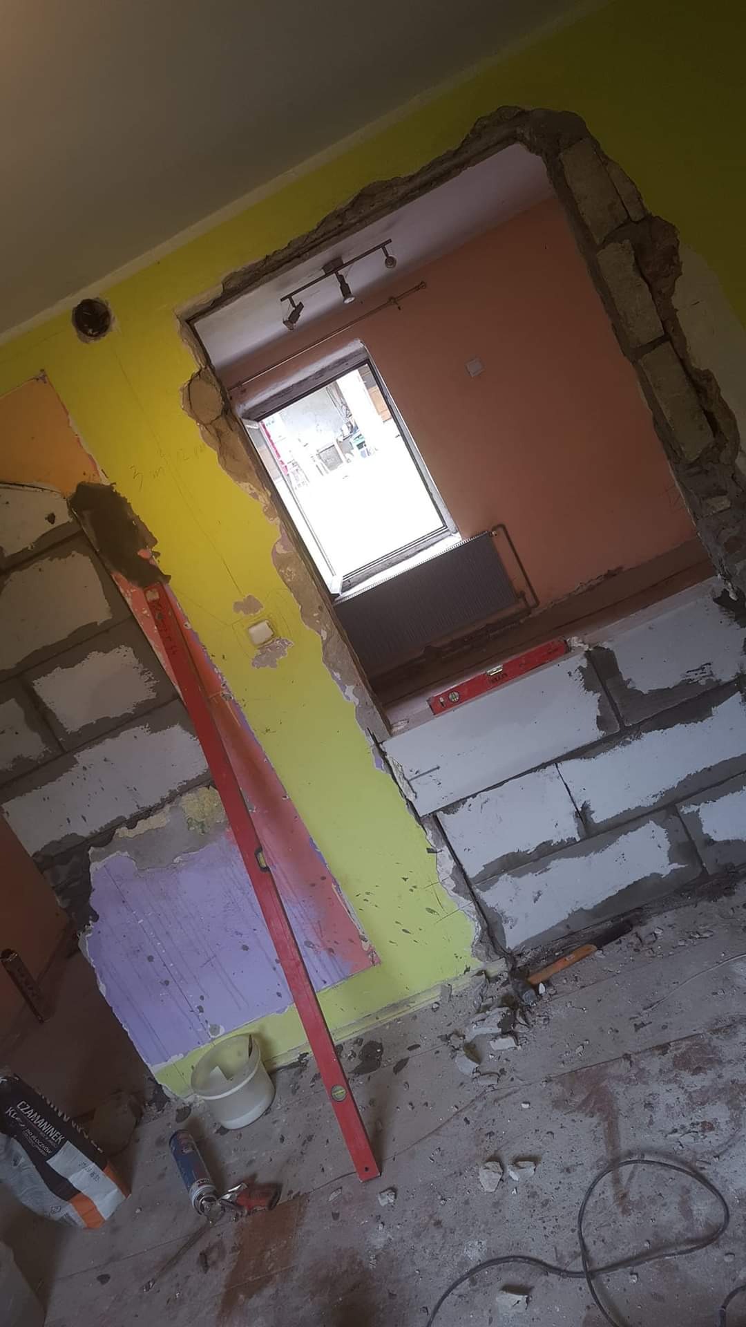 Wnętrze w trakcie prac remontowych: wznoszenie ściany z bloczków, widoczny poziomica, resztki starej farby na ścianach w odcieniach żółci i fioletu, oraz materiały budowlane na podłodze.