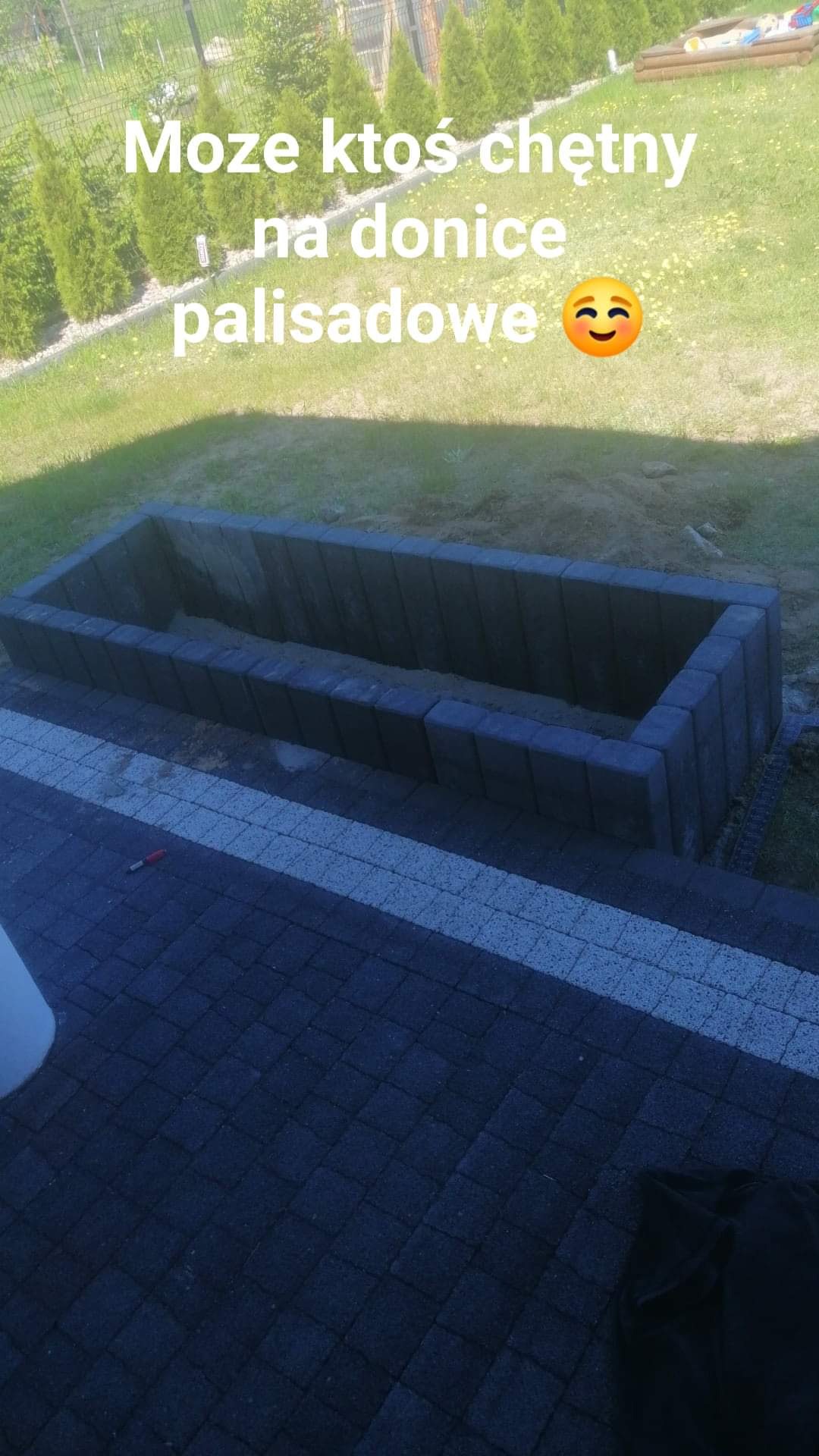 Murowana donica palisadowa z szarej kostki, wypełniona ziemią, na tle trawnika z tujami i kostki brukowej, widok z góry.