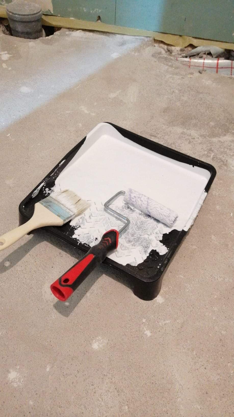 Kuветa z białą farbą, pędzel i wałek malarski z czerwoną rączką na podłodze w trakcie prac wykończeniowych w pomieszczeniu.