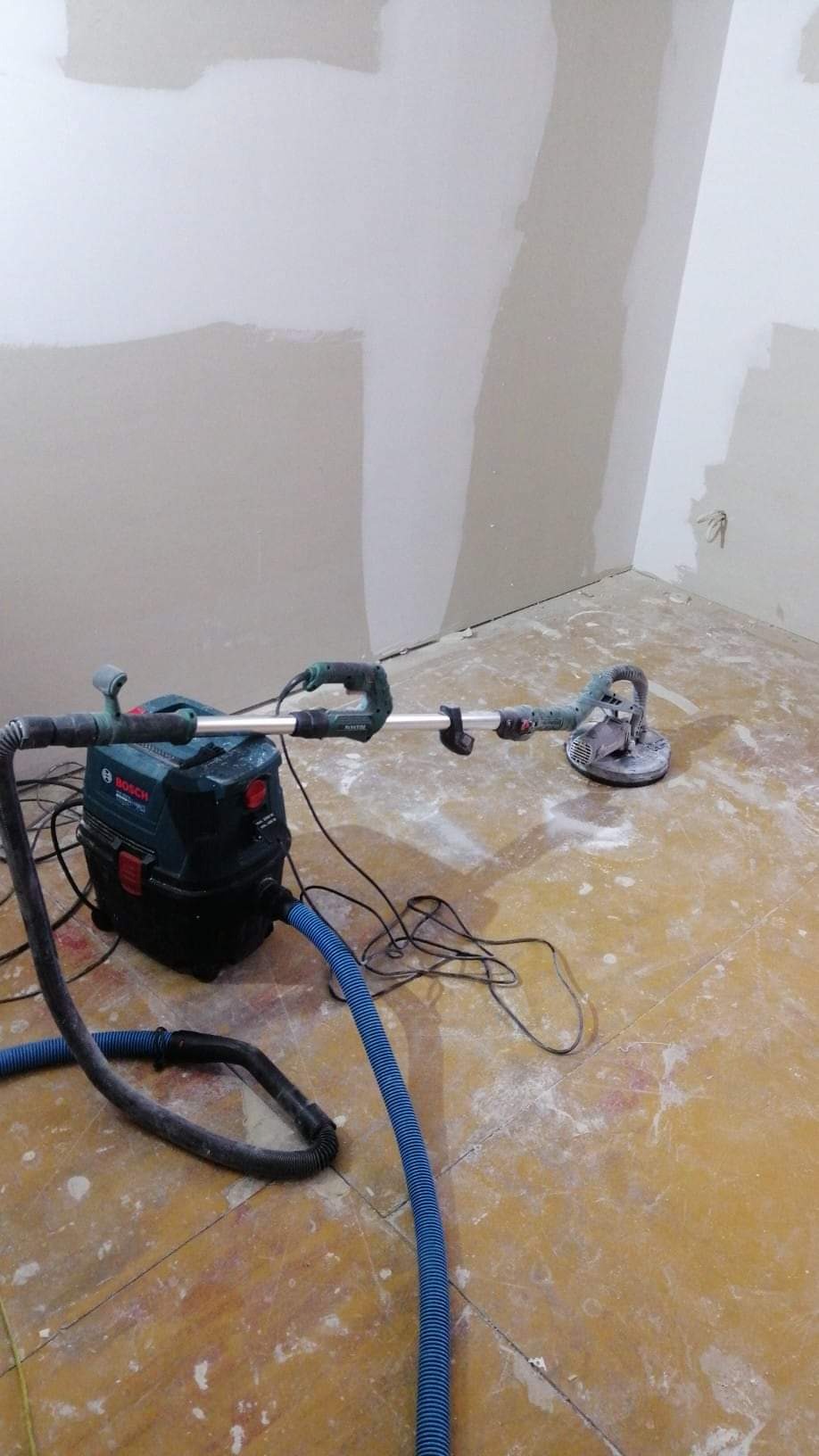 Szlifierka do gipsu z odkurzaczem przemysłowym Bosch podczas pracy na ścianie po szpachlowaniu, widoczne ślady gładzi na podłodze.