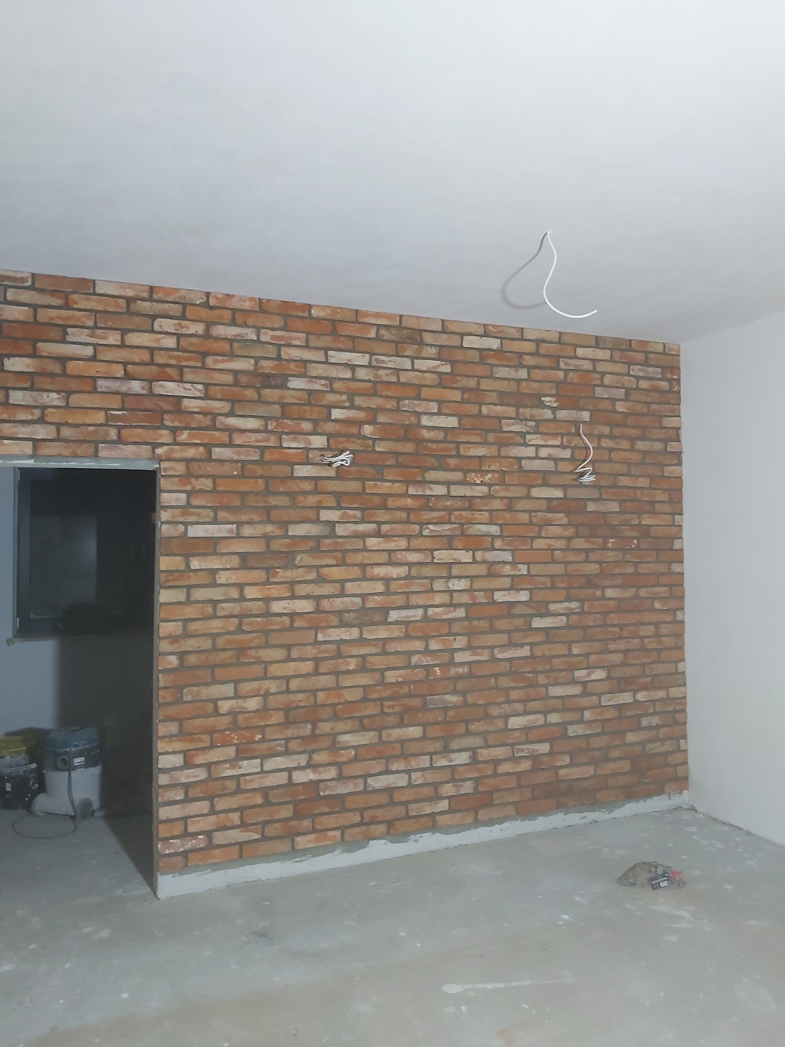 Ściana wyłożona dekoracyjną cegłą w pomieszczeniu w trakcie remontu, widoczne wystające kable elektryczne i odkurzacz przemysłowy w rogu pokoju.