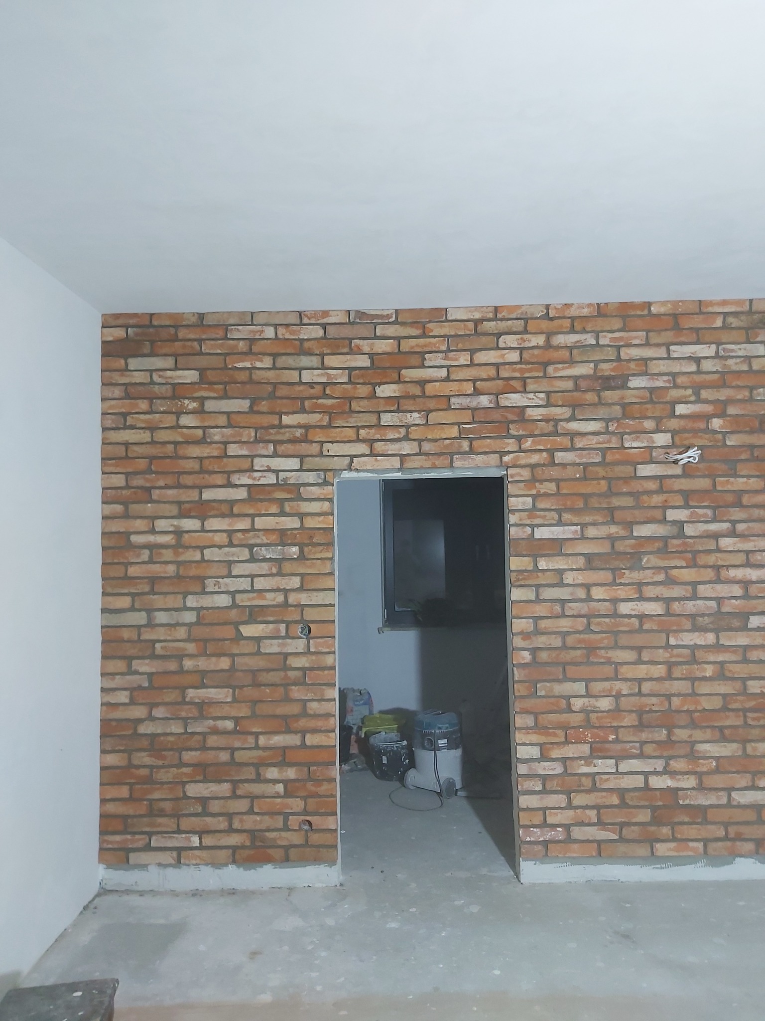 Ściana wykończona cegłą dekoracyjną w pomieszczeniu w trakcie remontu, widoczny odkurzacz przemysłowy w przejściu do kolejnego pokoju, niedokończone prace wykończeniowe przy podłodze.