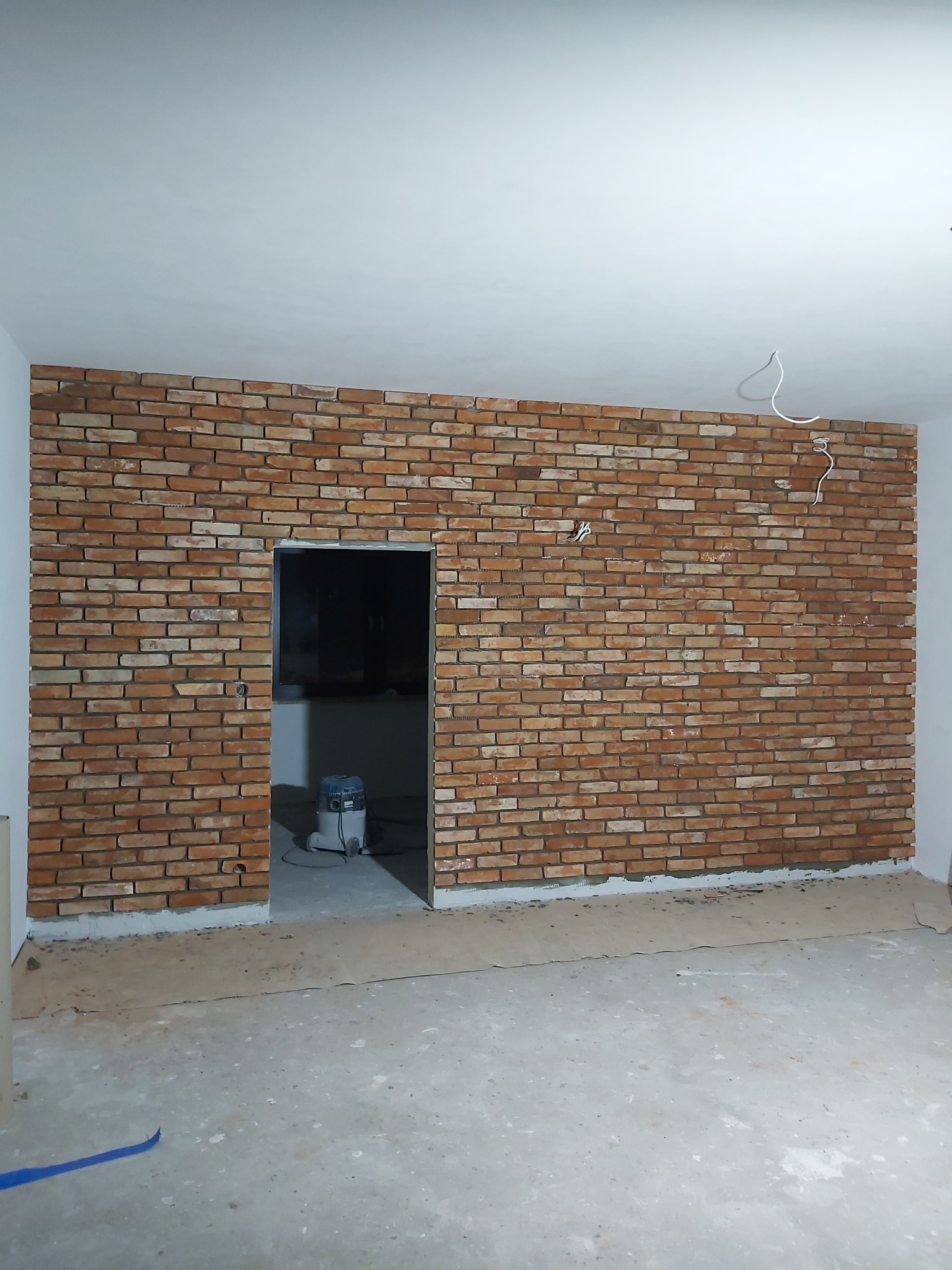 Ściana wykończona cegłą klinkierową w odcieniach czerwieni i brązu, widoczne przewody elektryczne wystające ze ściany, przygotowane do podłączenia oświetlenia, odkurzacz przemysłowy w przejściu...