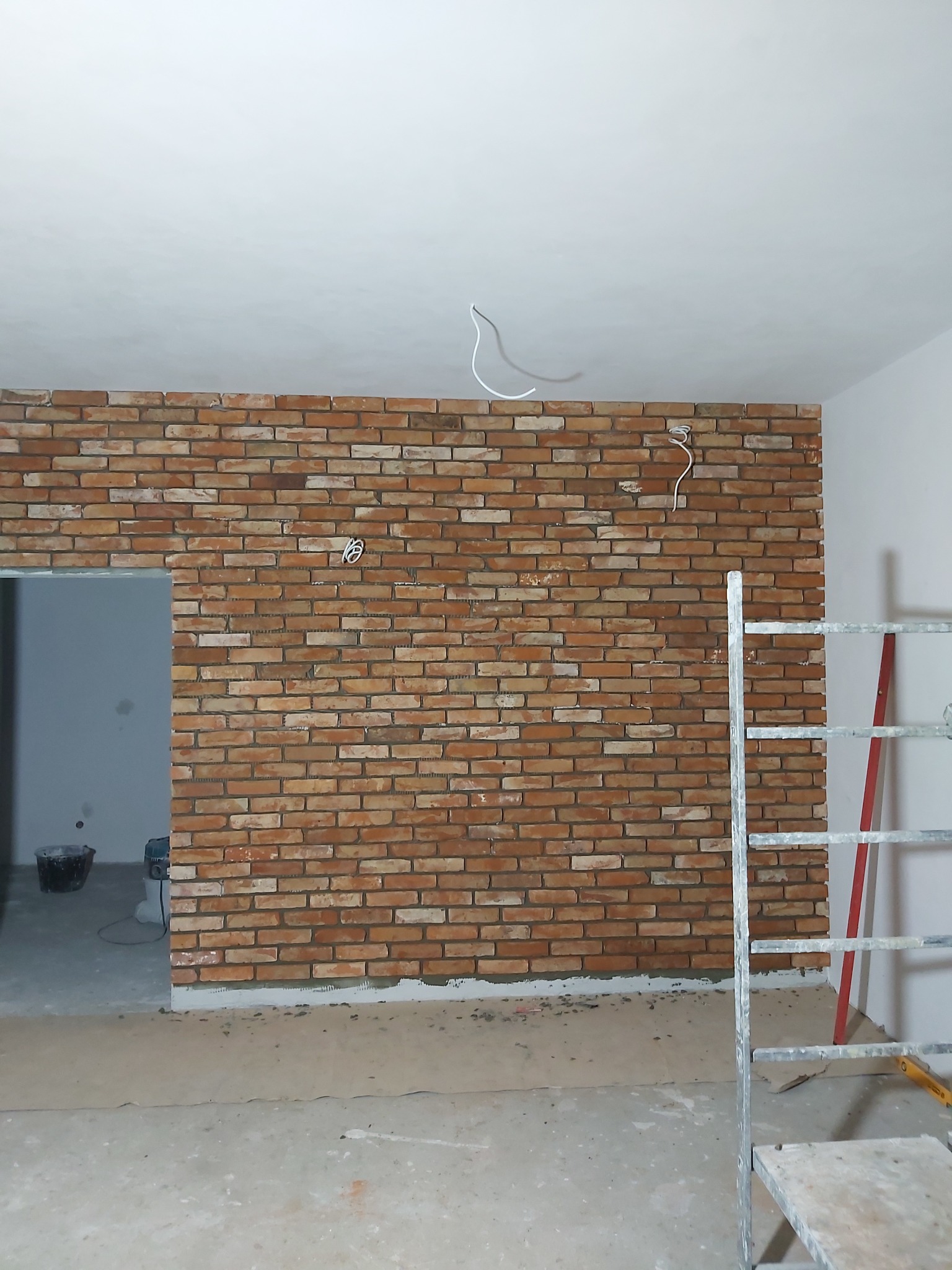 Ściana wewnętrzna wykończona cegłą dekoracyjną w odcieniach czerwieni i brązu, widoczne przewody elektryczne zwisające z sufitu, drabina aluminiowa w rogu, podłoga częściowo zabezpieczona kartonem.