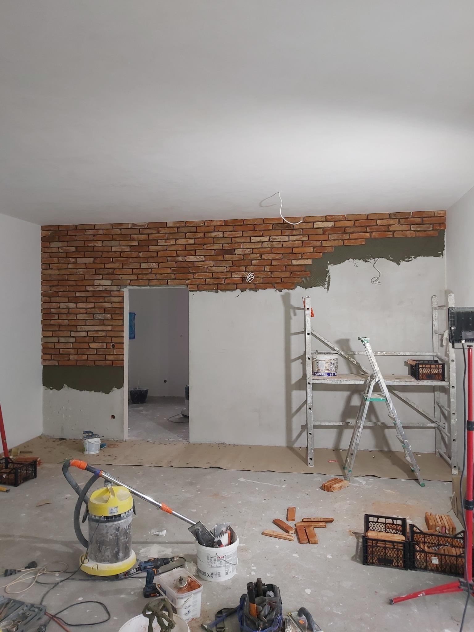 Montaż okładziny z cegły klinkierowej na ścianie wewnętrznej, widoczny fragment ściany z nałożonym klejem, drabina, narzędzia i materiały budowlane na podłodze w trakcie prac wykończeniowych.