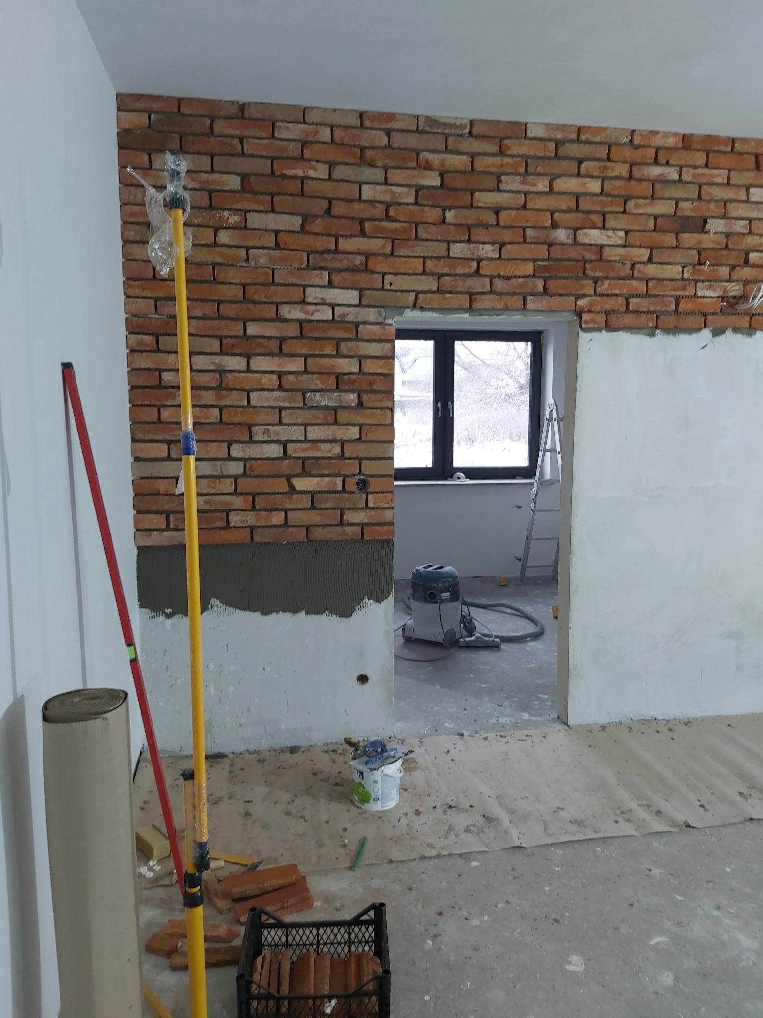 Wnętrze w trakcie prac wykończeniowych, fragment ściany z cegły klinkierowej, częściowo ułożonej, widoczne narzędzia, poziomica, odkurzacz przemysłowy w tle, przygotowane cegły w koszyku na podłodze.