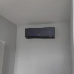 Czarny klimatyzator marki Gree zamontowany na szarej ścianie w pomieszczeniu, widoczny krzyż na ścianie nad drzwiami i plakaty w ramkach po prawej stronie.