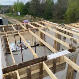 Wznoszenie ścian z bloczków, widoczne drewniane szalunki i zbrojenie, w tle zieleń.