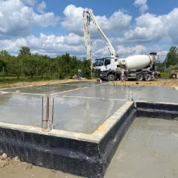 Wylany fundament z hydroizolacją, widoczny betonowóz z pompą do betonu na tle zieleni i błękitnego nieba.