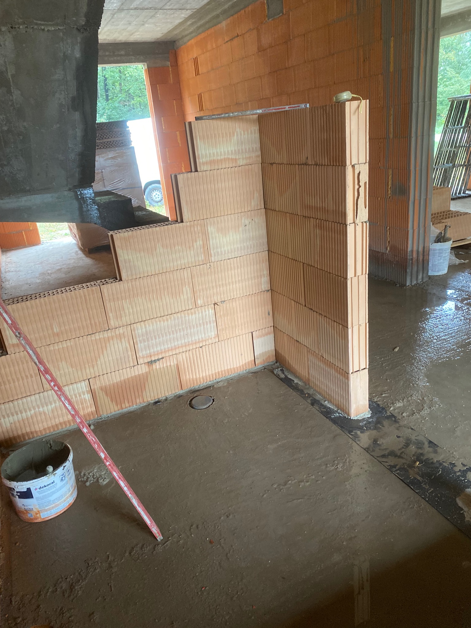 Wnętrze w trakcie budowy z widocznymi ścianami z czerwonej cegły, mokrą posadzką i wiadrem z zaprawą, z widocznym poziomem na ścianie.