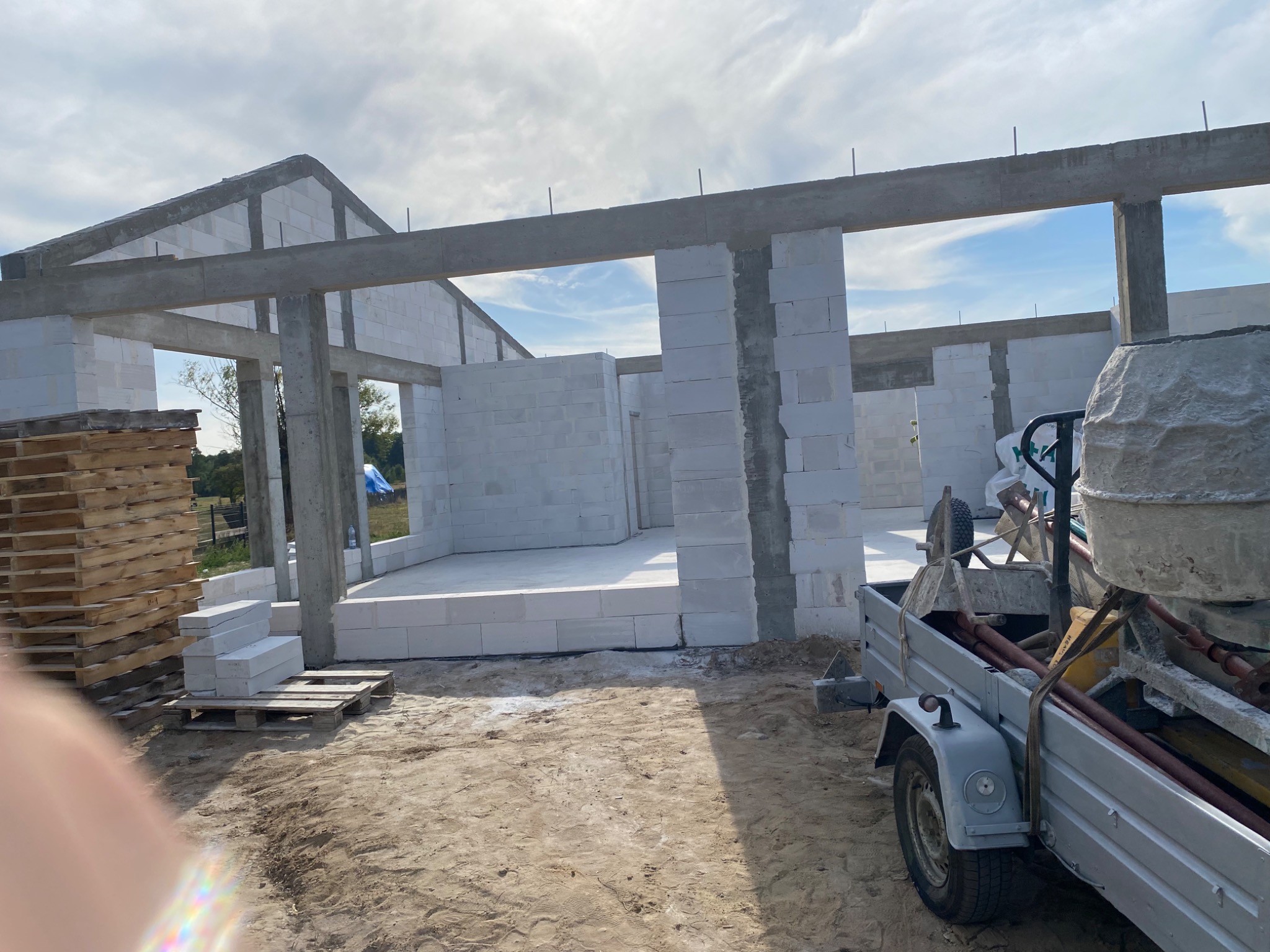 Surowy stan domu z betonu komórkowego w trakcie budowy, widoczne ściany, strop i przygotowane palety z bloczkami, na pierwszym planie przyczepka z betoniarką.